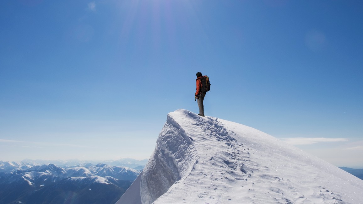 Ein Bergsteiger steht vor tiefblauem Himmel auf einer schneebedeckten Bergspitze und schaut in die Ferne.