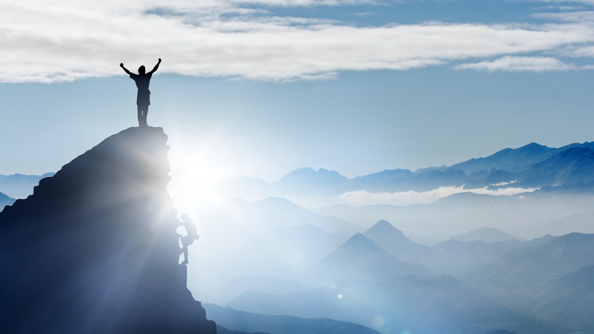 Ein Mann steht im Sonnenaufgang mit jubelnd nach oben ausgestreckten Armen auf einer Bergspitze vor einem Gebirgspanorama.