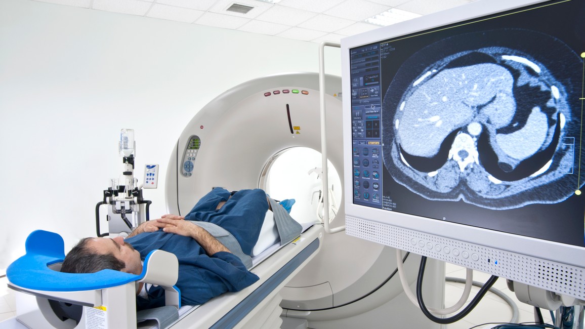 Ein Patient liegt auf dem Rücken auf der ausgefahrenen Patientenliege vor der Untersuchungsröhre eines MRT. Rechts daneben zeigt ein Monitor MRT-Bilder eines Gehirns.