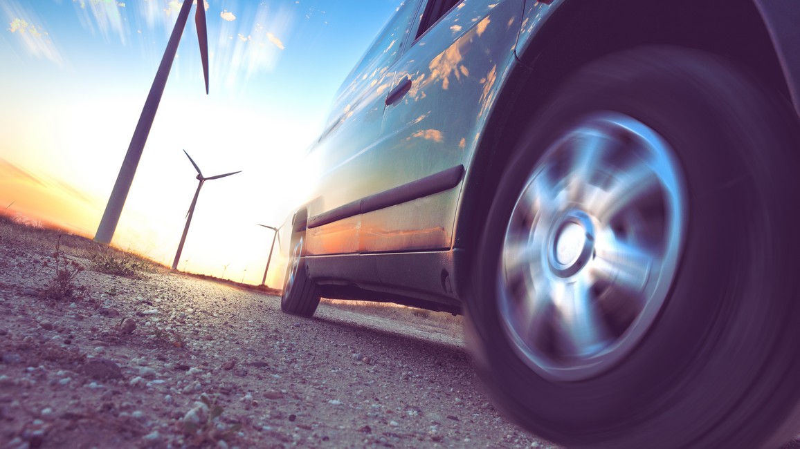 Im Vordergrund fährt ein Auto, das nur ausschnittsweise zu sehen ist, auf einer Straße. Im Hintergrund sind links mehrere Windturbinen im Abendlicht zu sehen.