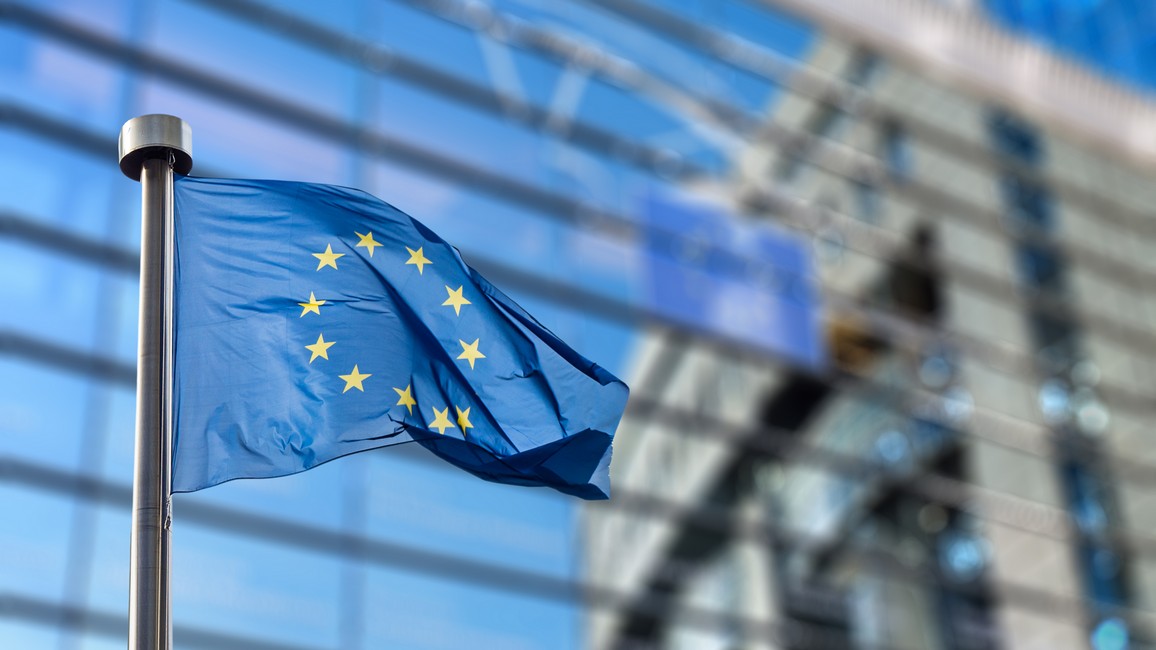 Eine EU-Flagge flattert vor dem Berlaymont-Gebäude in Brüssel, dem Sitz der Europäischen Kommission. Das Gebäude ist verschwommen ausschnittsweise im Hintergrund zu sehen.