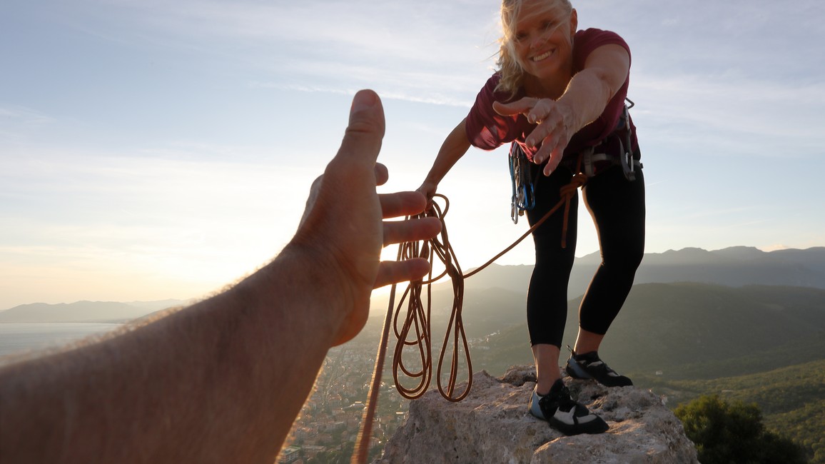 Eine Frau reicht bei einer Klettertour im Gebirge einem angeseilten Mann die Hand, um ihn beim Aufstieg zu unterstützen.