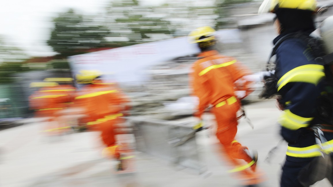 Das Bild zeigt mit Verzerrungen mehrere Rettungskräfte im Noteinsatz, die von rechts nach links laufen. Zwei der Rettungskräfte tragen eine Bahre zwischen sich.